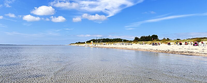 Flaches, klares Meerwasser vor einem Sandstrand mit Strandkörben unter blauem Himmel mit weißen Wolken