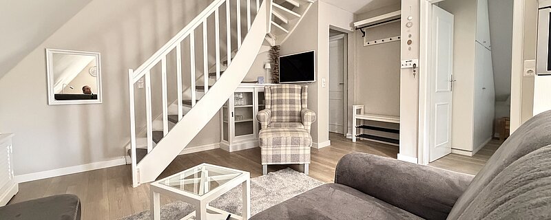 Wohnzimmer mit grauem Sofa, weißem Glastisch, kariertem Sessel und weißer Treppe ins Dachgeschoss