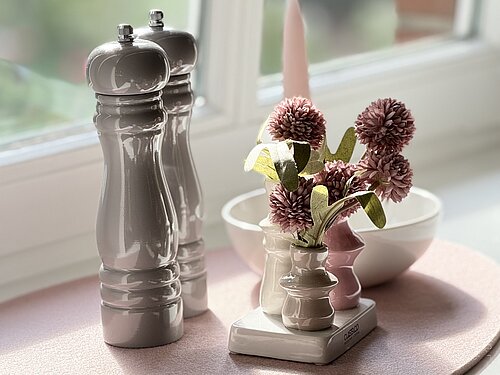 Zwei Gewürzmühlen, eine kleine Blumenvase und eine rosa Kerze auf einer rosa Unterlage am Küchenfenster