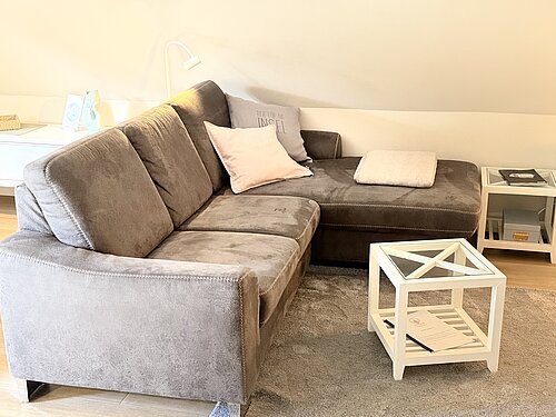 Graues Ecksofa mit hellen Kissen und gefalteter Decke unter der Dachschräge, davor ein weißer Glastisch