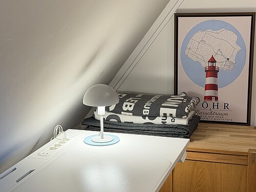 Arbeitsplatz unter der Dachschräge mit weißem Schreibtisch, kleiner Lampe und einem Föhr-Poster auf der Holztruhe