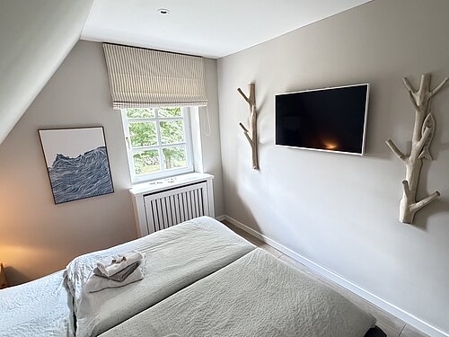 Doppelbett mit hellen Decken und gefalteten Handtüchern; gegenüber hängen ein Fernseher und Garderoben aus Ästen