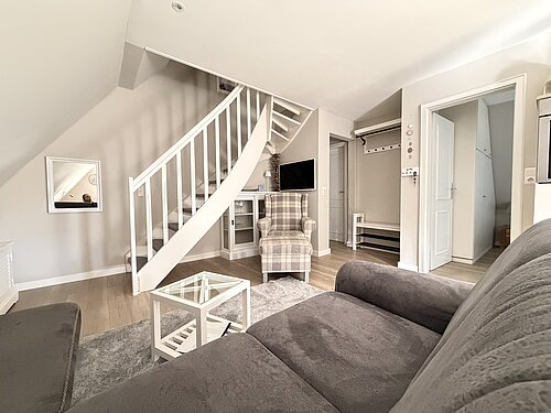 Wohnzimmer mit grauem Sofa, weißem Glastisch, kariertem Sessel und weißer Treppe ins Dachgeschoss