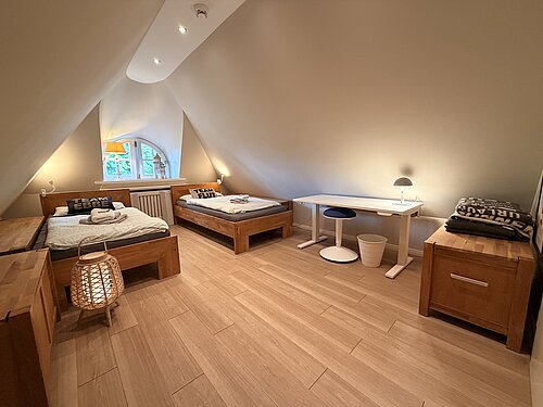 Schlaf- und Arbeitszimmer unter steilen Dachschrägen mit zwei Einzelbetten, weißem Schreibtisch und Holztruhen