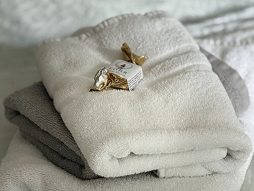 Nahaufnahme von gefalteten weißen und grauen Handtüchern mit einem goldfarben verpackten Bonbon auf dem Bett