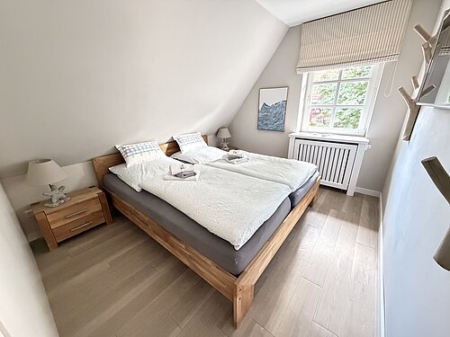Schlafzimmer unter der Dachschräge mit hölzernem Doppelbett, zwei Nachttischen und einem Fenster mit gestreiftem Raffrollo